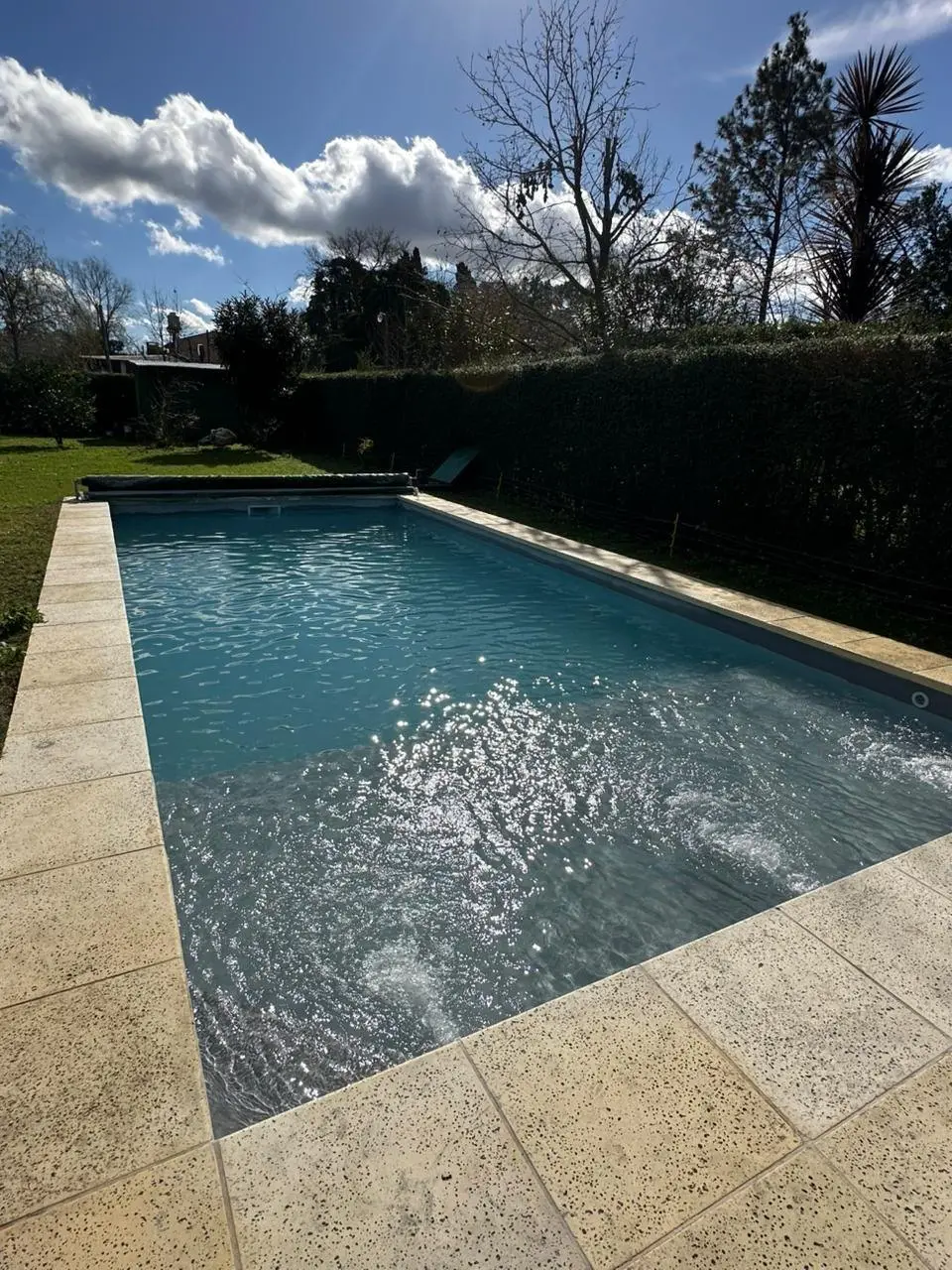 Piscina rectangular con solado de piedra clara, rodeada de un cerco verde y árboles