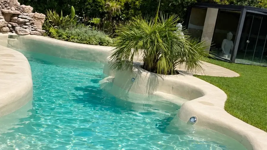 Piscina tipo laguna con agua turquesa, terminación de arena e isla con palmera en un jardín de Nordelta