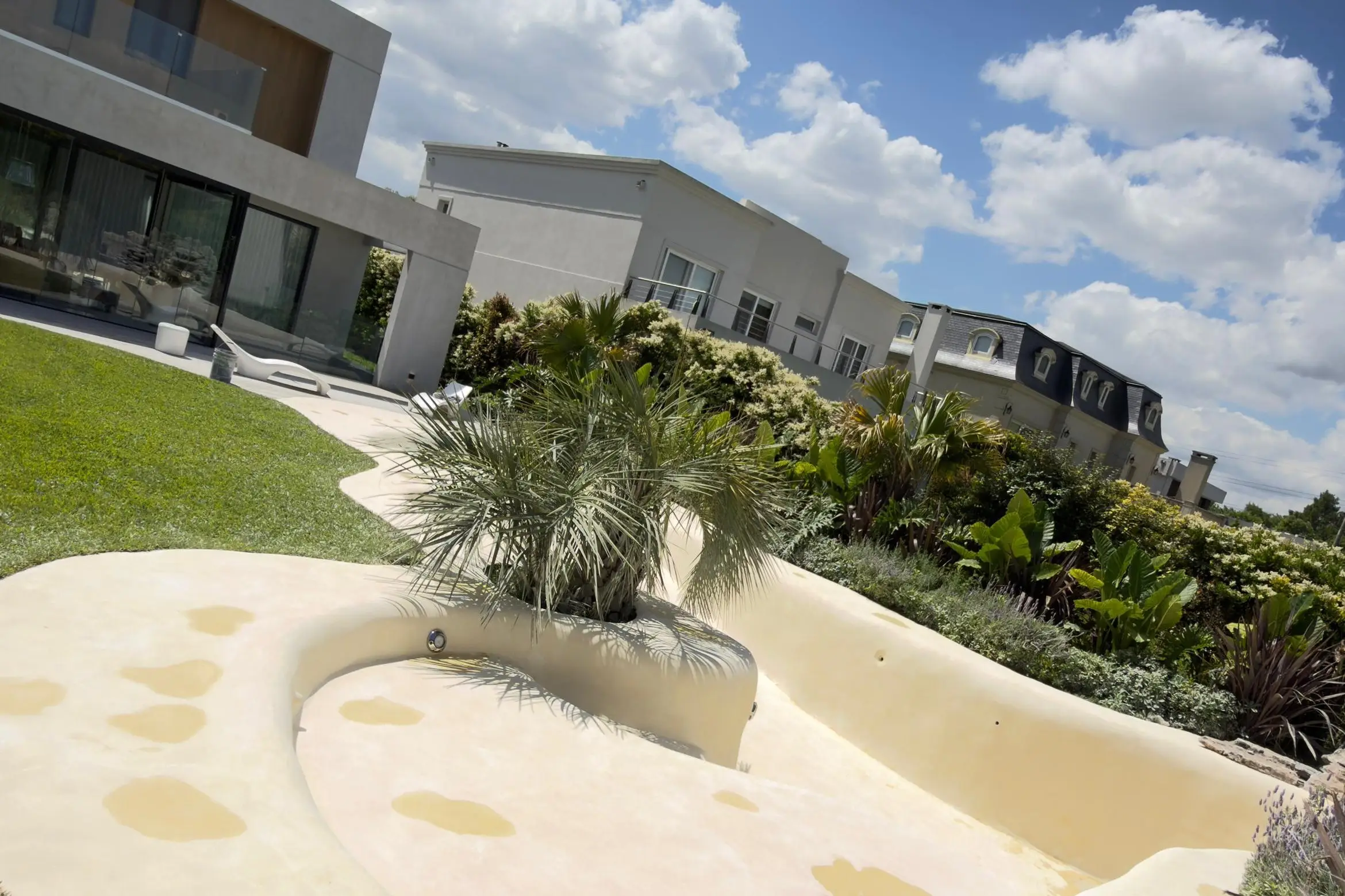 Piscina de arena de formas curvas con isla de palmeras frente a una casa moderna