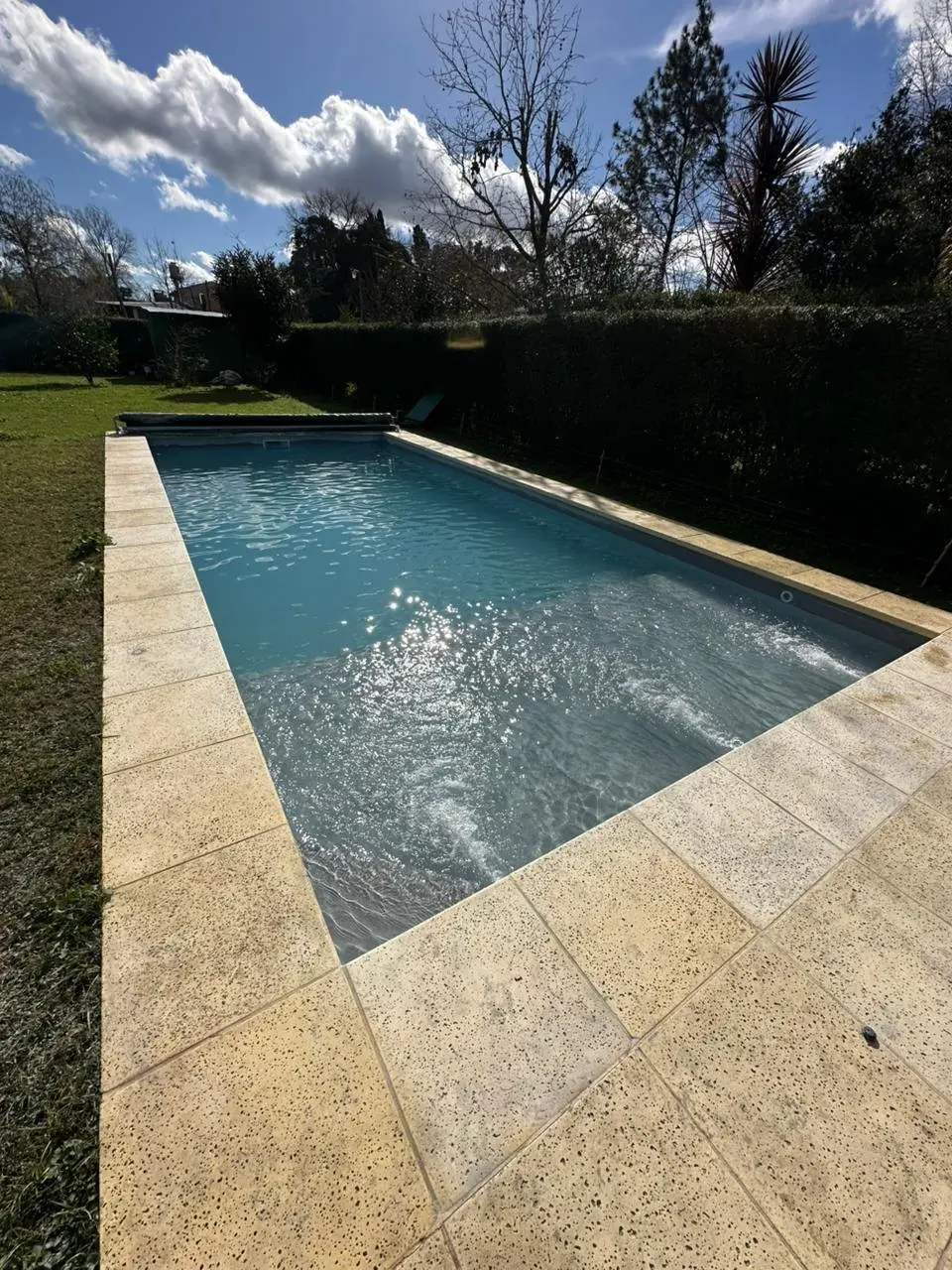 Solado de piedra clara alrededor de una piscina rectangular, con un cerco verde de fondo
